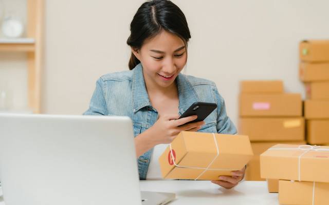 A sourcing agent checking a parcel against an order on her phone, next to a laptop and boxes